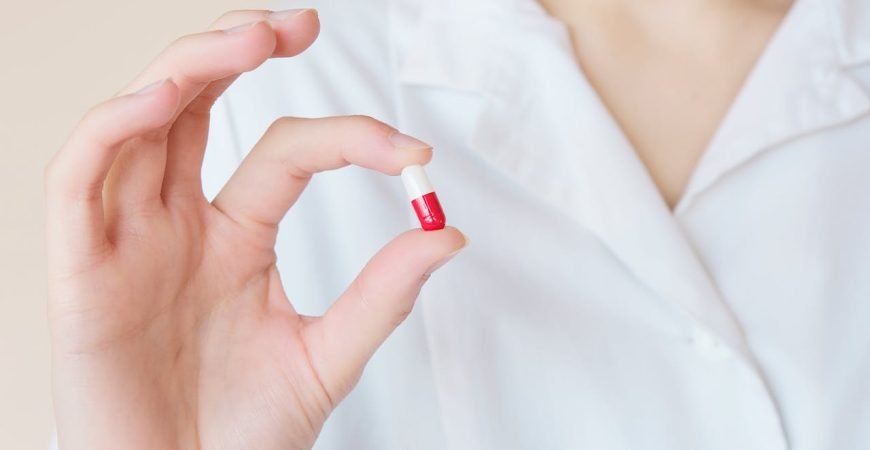 Persona con bata blanca mostrando una cápsula roja y blanca, representando la profesión farmacéutica